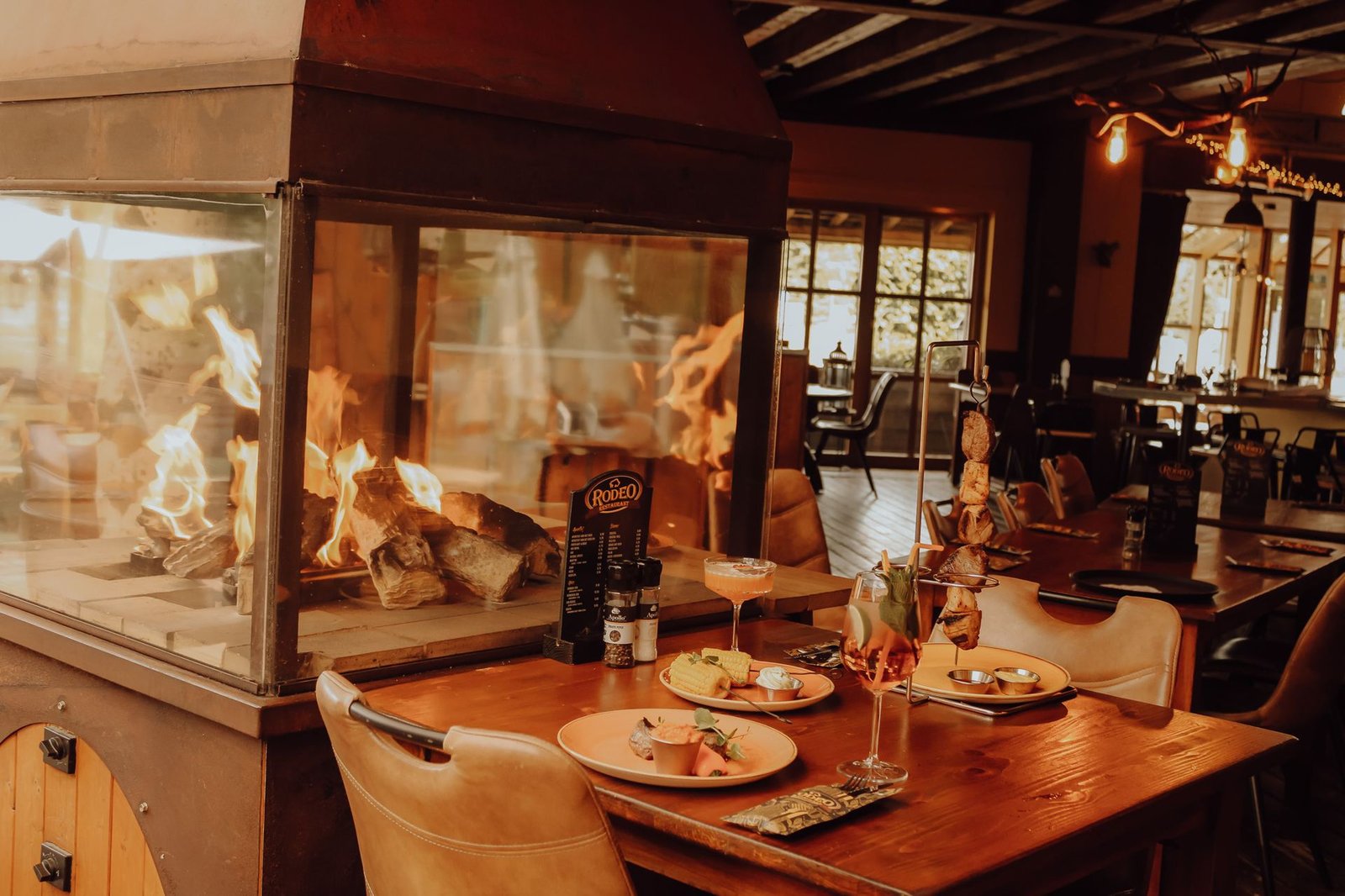 Gedekte tafel met cocktails en een vleesspies, met het open haardvuur van Saloon Rodeo op de achtergrond